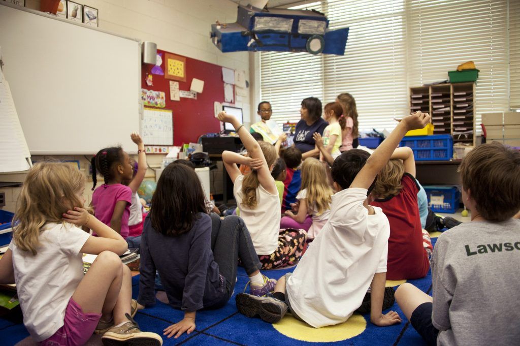 niños y niñas sentados en el suelo en una clase levantan la mano